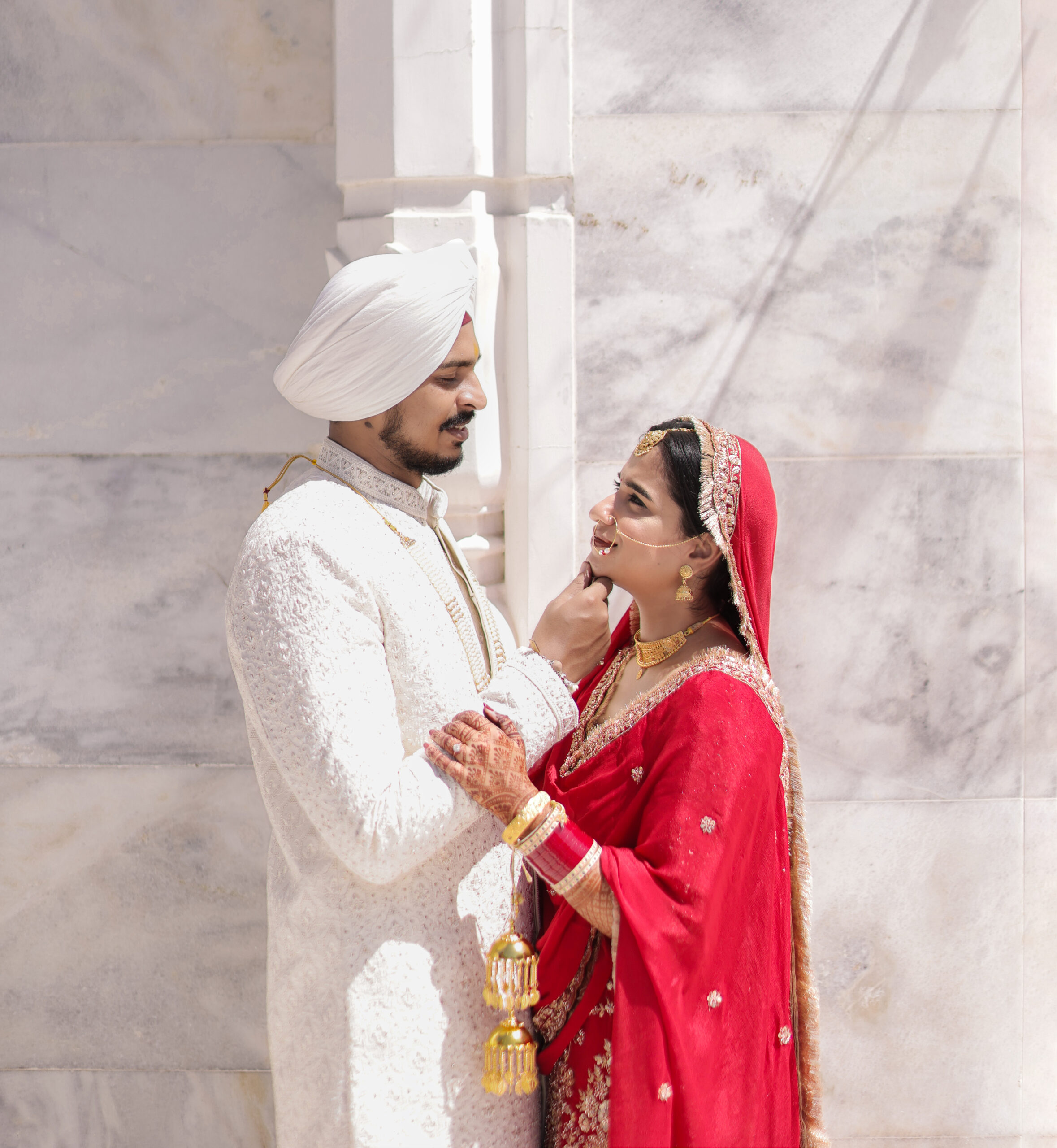 A sunlit outdoor portrait of a Sikh bride and groom standing closely together in front of a white marble wall. The groom, wearing a white embroidered sherwani and turban, gently touches the bride's chin. The smiling bride wears a vibrant red outfit with gold embroidery, traditional red chooda bangles, golden kalire, and heavy gold bridal jewelry.