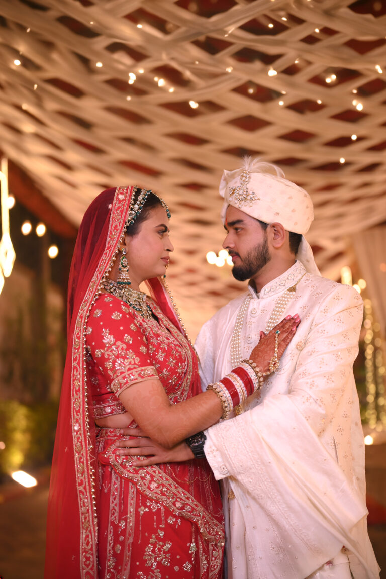 Royal Indian Wedding Photography – Elegant portrait of a bride in a red bridal lehenga and groom in an ivory sherwani sharing a romantic moment under beautifully decorated wedding lights.