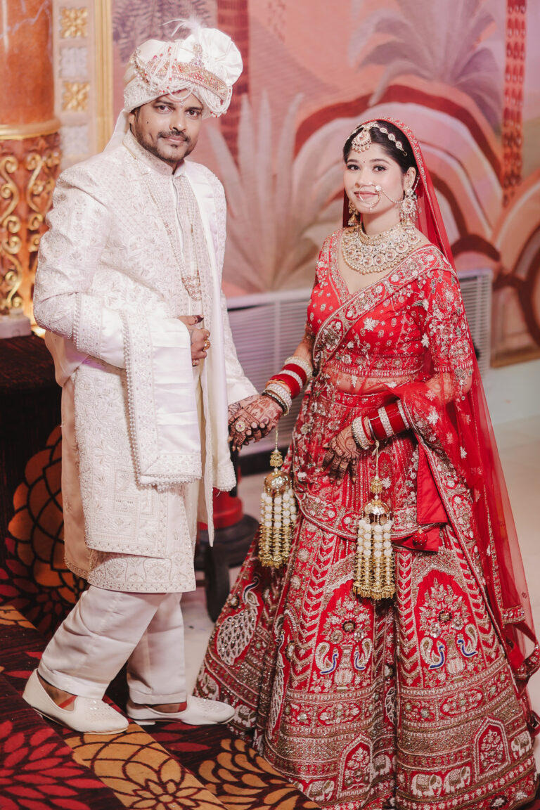 A full-length portrait of a smiling Indian bride and groom holding hands. The bride wears a heavily embroidered red bridal lehenga with traditional kalire and jewelry. The groom wears an intricately embroidered ivory sherwani with a matching turban and mojari shoes, standing against a painted pastel mural backdrop.