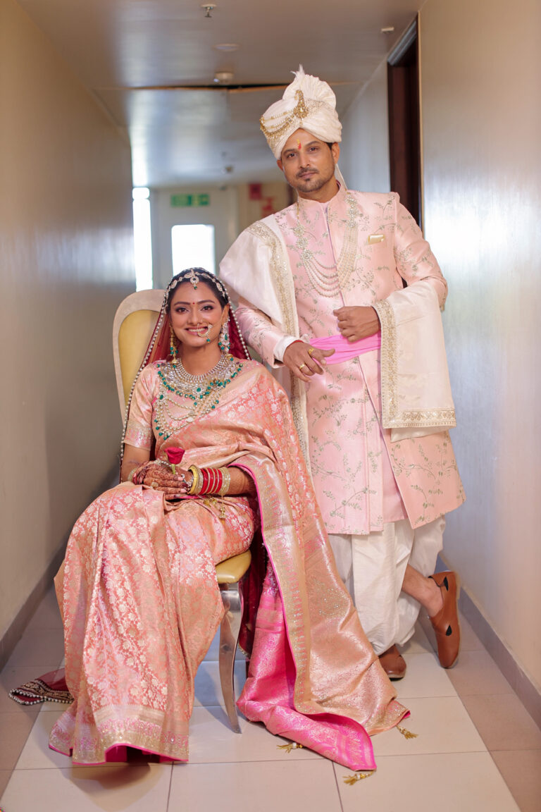 A smiling Indian couple in matching blush pink traditional wedding attire. The bride is seated wearing a heavily embroidered pink silk saree and jewelry, while the groom stands beside her in a pink sherwani, pearl mala, and white turban, posing in a well-lit modern corridor.