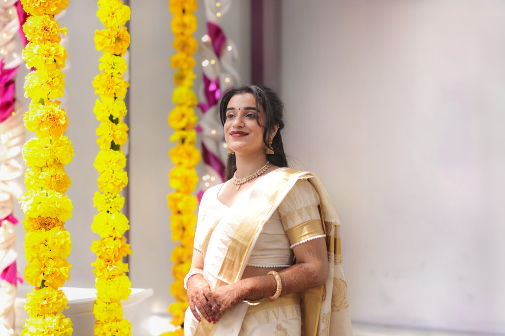 An indoor portrait of a smiling woman standing gracefully next to vibrant hanging strands of yellow marigold flowers. She wears a classic white and gold traditional saree, a delicate necklace, earrings, and mehendi on her hands.