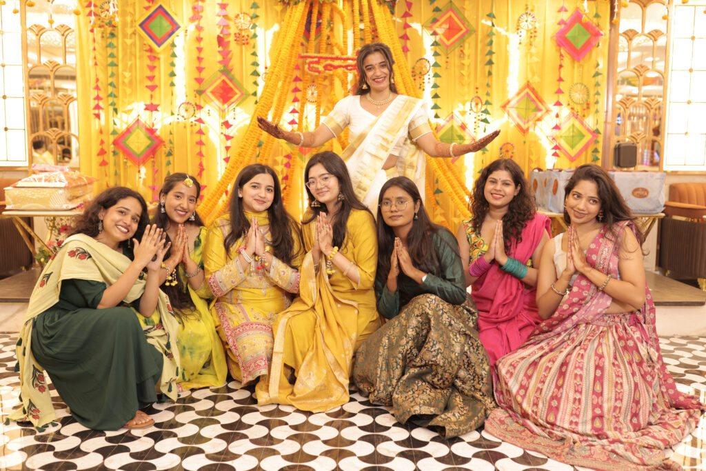 An indoor group portrait of a woman standing at the center with her arms spread in white and gold traditional attire, surrounded by a group of smiling women sitting below her on a checkered floor with hands held in a Namaste gesture. They wear colorful traditional outfits against a backdrop of yellow drapes and marigold flowers.