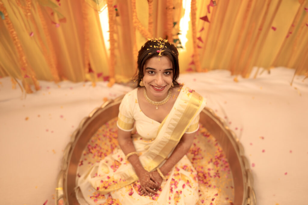 An elevated, high-angle indoor portrait of a smiling bride sitting inside a golden, lotus-shaped basin filled with flower petals. She wears a traditional white and gold saree, a delicate necklace, and has turmeric and flower petals gently scattered on her hair and shoulders, set against a bright yellow and white backdrop.
