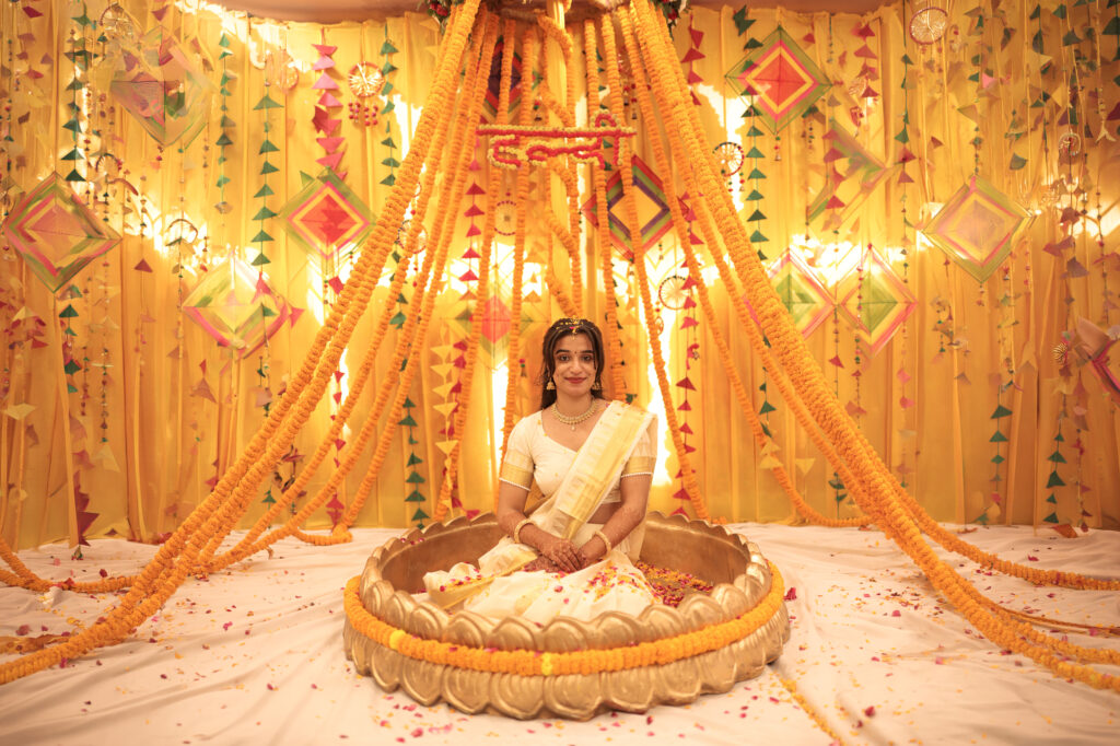An indoor portrait of a smiling bride sitting gracefully inside a flower-filled lotus basin during her Haldi ceremony. She wears a traditional white and gold saree, seated beneath an elaborate canopy constructed from cascading strings of bright yellow marigold flowers and colorful hanging kite decorations.