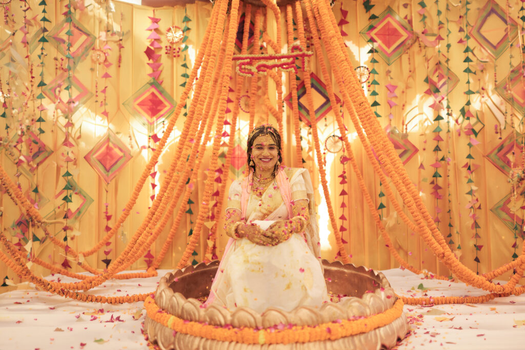 An indoor portrait of a smiling bride sitting inside a decorative lotus-shaped basin during her Haldi ceremony. She wears a white and gold outfit smeared with turmeric, seated beneath a grand canopy of marigold flower garlands, colorful hanging paper kites, and geometric backdrops.