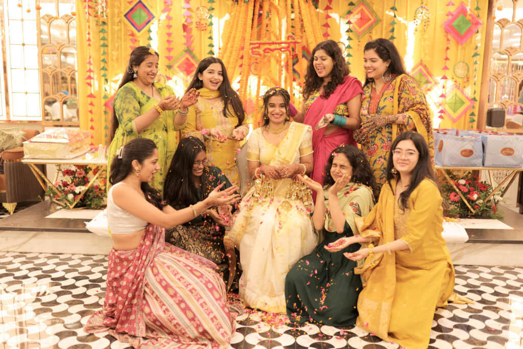 An indoor group portrait of a bride seated centrally in white and gold traditional attire, smiling happily as friends and family surround her, playfully showering her with flower petals. The guests wear colorful ethnic outfits and yellow traditional wear against a vibrant marigold-decorated background.