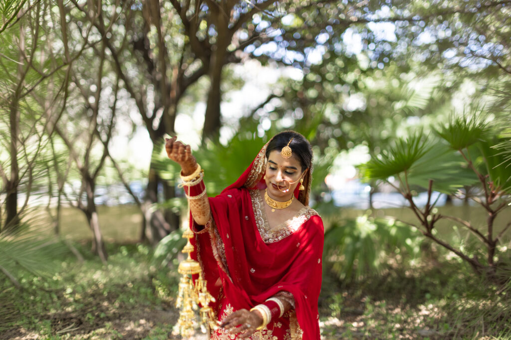 An outdoor portrait of a bride standing outdoors amongst lush green trees and foliage, wearing a vibrant red traditional outfit with a veil. She smiles down gently while holding up her traditional bridal kalire ornaments, adorned with a gold necklace, maang tikka, and nose ring.