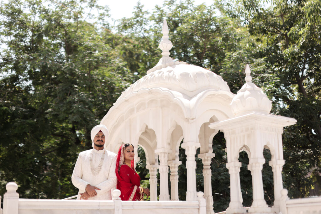 An outdoor portrait of a couple standing near an ornate white marble pavilion (chhatri) surrounded by lush green trees. The man wears a classic cream sherwani with a matching turban and a beaded necklace, while the woman wears a vibrant red traditional outfit with a veil and gold jewelry.