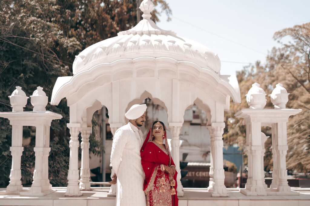 An outdoor portrait of a newlywed couple posing inside an ornate white marble pavilion (chhatri) at a Gurudwara, surrounded by green trees. The groom wears a cream sherwani with a matching turban, while the bride looks up at him lovingly, wearing a vibrant red traditional outfit with a veil and gold jewelry.