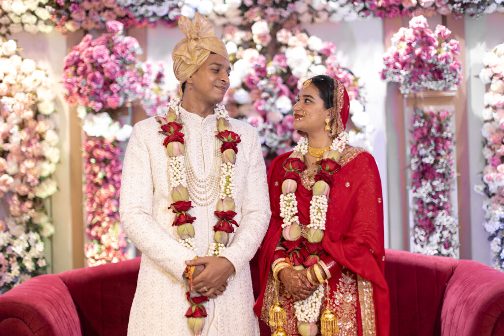 An indoor portrait of a newlywed couple standing together and looking lovingly at each other. The groom wears a white embroidered sherwani, a matching turban, pearl necklaces, and a floral garland, while the bride shines in a traditional red lehenga, veil, gold jewelry, and a matching floral garland against a backdrop of lush pink and white floral arrangements.