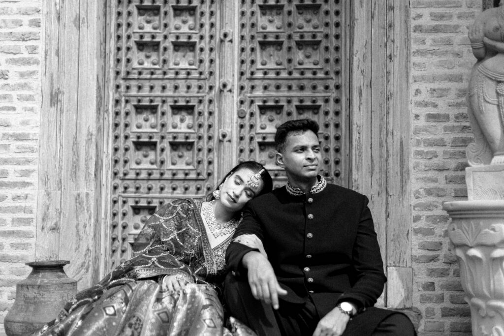 A black-and-white outdoor portrait of a couple posing against an intricately carved wooden door and brick wall. The groom wears a dark traditional bandhgala jacket, while the bride rests her head gently on his shoulder, wearing an embellished lehenga and traditional jewelry.
