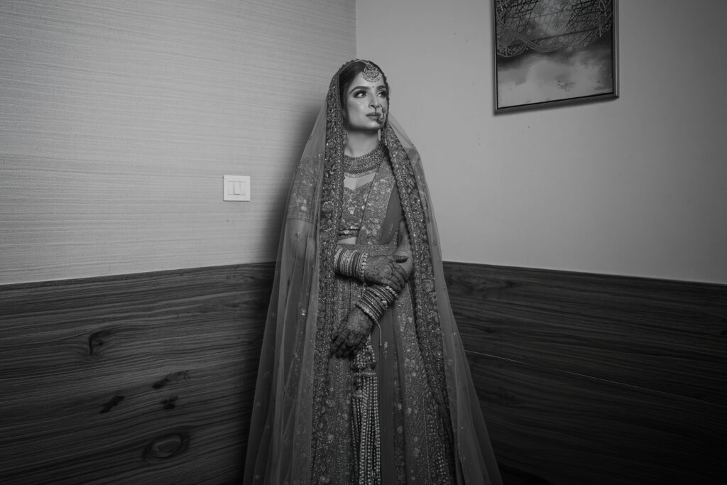 A black-and-white indoor portrait of a bride standing gracefully against a wood-paneled wall, wearing an ornate heavily-embellished lehenga, bridal veil, choker necklace, and traditional bangles.