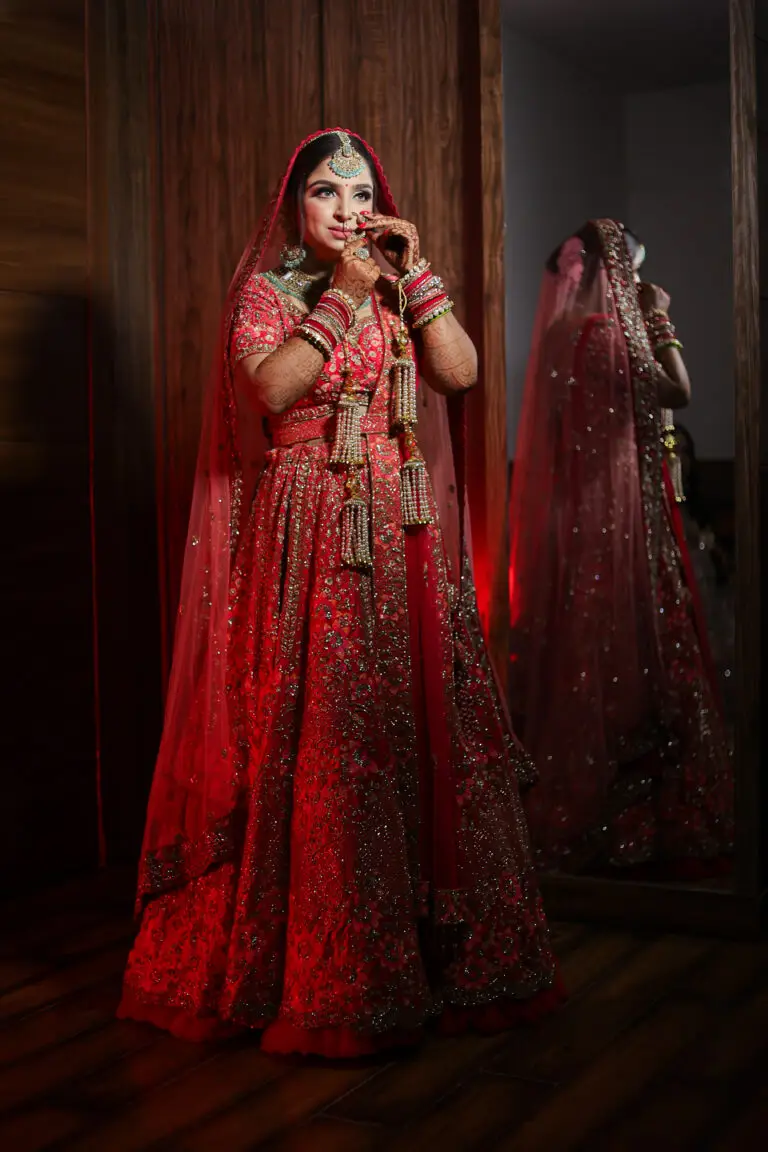 A full-length, low-light portrait of an Indian bride adjusting her nath (nose ring) in front of a tall mirror. She is wearing a heavily embroidered red bridal lehenga and heavy kundan jewelry. Her reflection is visible in the mirror, illuminated by dramatic, warm red lighting.