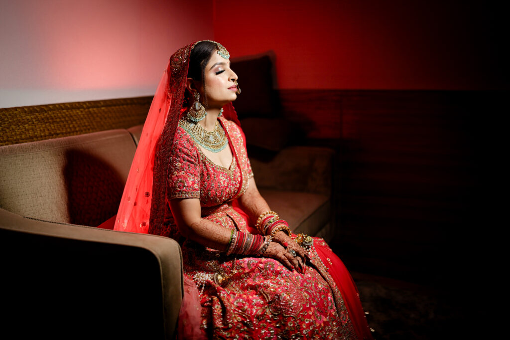 An indoor portrait of a bride sitting gracefully on a sofa with her eyes closed peacefully. She wears a heavily-embellished red lehenga and veil, complemented by a mint-green and gold statement choker necklace and earrings, set against a warm red-lit background.