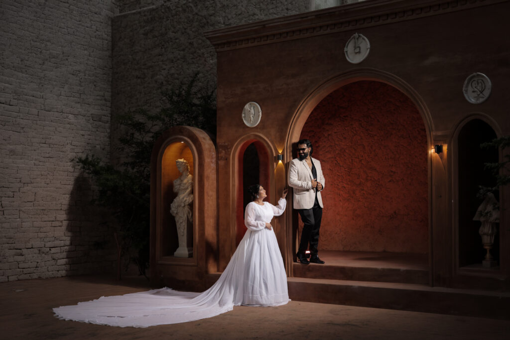 A wide indoor portrait of a couple posing against a warm, terracotta-toned architectural backdrop featuring arched niches, classical statues, and textured walls. The woman wears a flowing white gown with a long train, while the man stands in an arched doorway wearing a light patterned blazer and dark trousers.