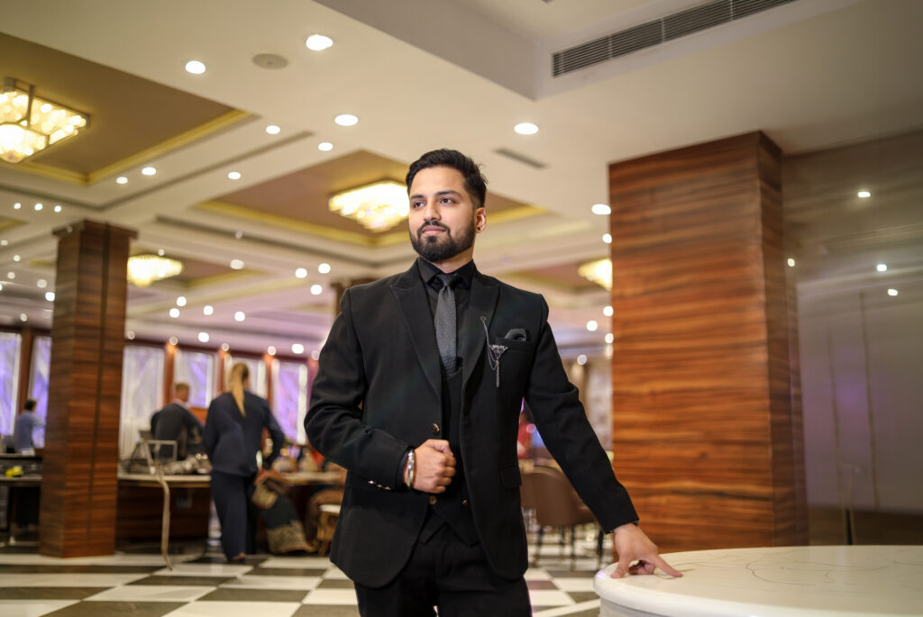 An indoor portrait of a man standing inside a luxurious banquet hall with a checkered floor, warm ceiling lights, and wood-paneled pillars. He wears a sharp black three-piece suit with a black shirt, tie, and a lapel accessory, resting one hand lightly on a round table while looking off to the side.