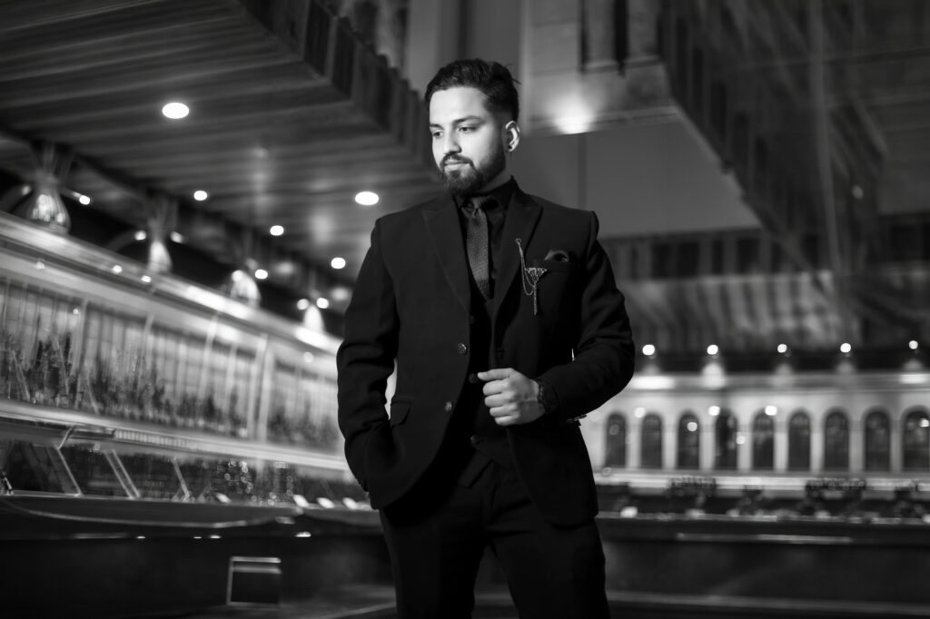 A striking black-and-white portrait of a man standing inside a grand, warmly lit hall with reflections and modern architecture. He wears a tailored three-piece suit, a dark tie, and a lapel pin accessory, posing confidently with one hand in his pocket.