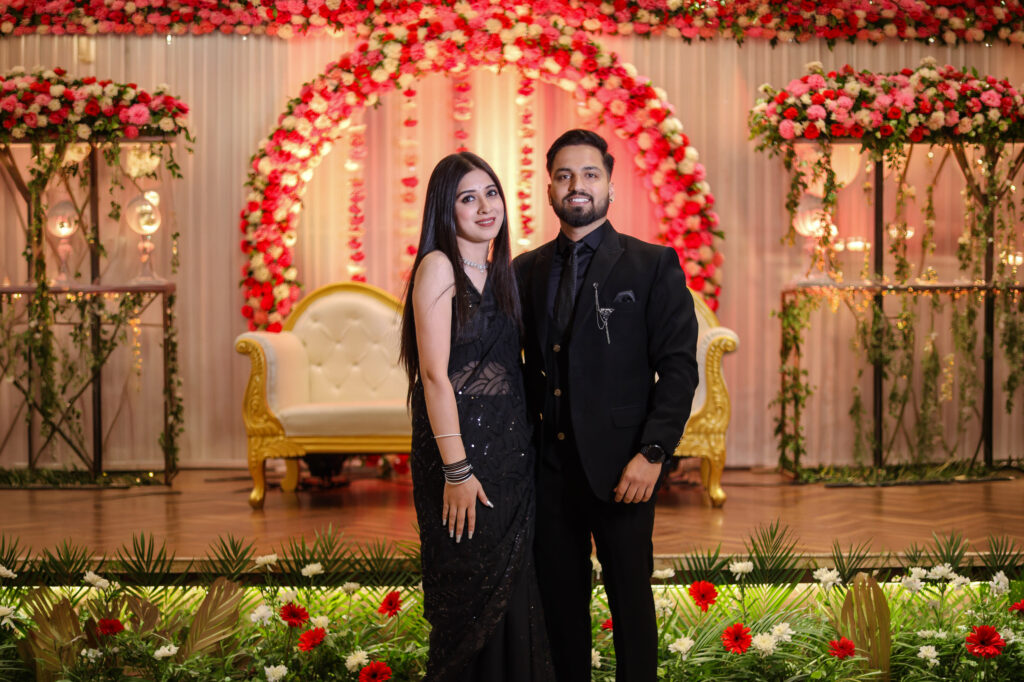 An indoor portrait of a couple standing side-by-side in front of a floral arch backdrop decorated with pink and white flowers. The woman wears a sparkling black sheer saree with silver detailing and a necklace, and the man wears a matching black suit, shirt, and tie with a lapel chain accessory.