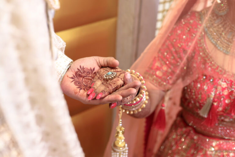 A close-up indoor shot of a couple during an engagement or ring ceremony. A hand with mehendi and red nail polish holds out a delicate ring with a central gemstone, while traditional wedding attire, bangles, and kalire are softly visible.