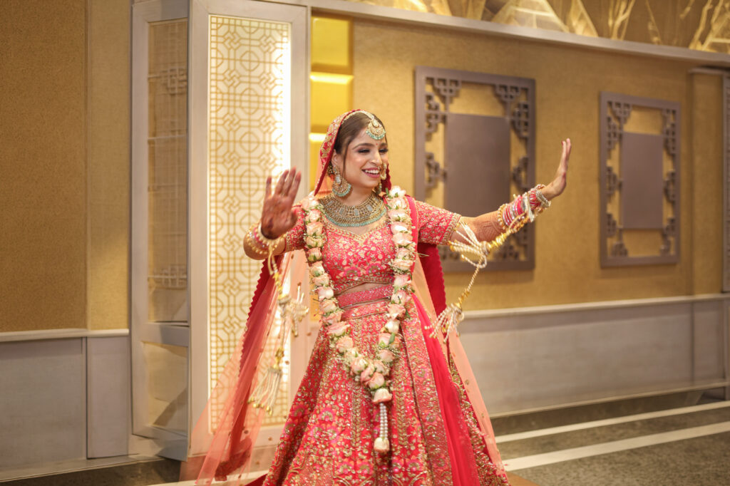 An indoor portrait of a smiling bride dancing gracefully with her arms raised and hands open. She wears a vibrant pink and gold embroidered lehenga, a matching veil, traditional statement jewelry, a floral garland, and bridal kalire.