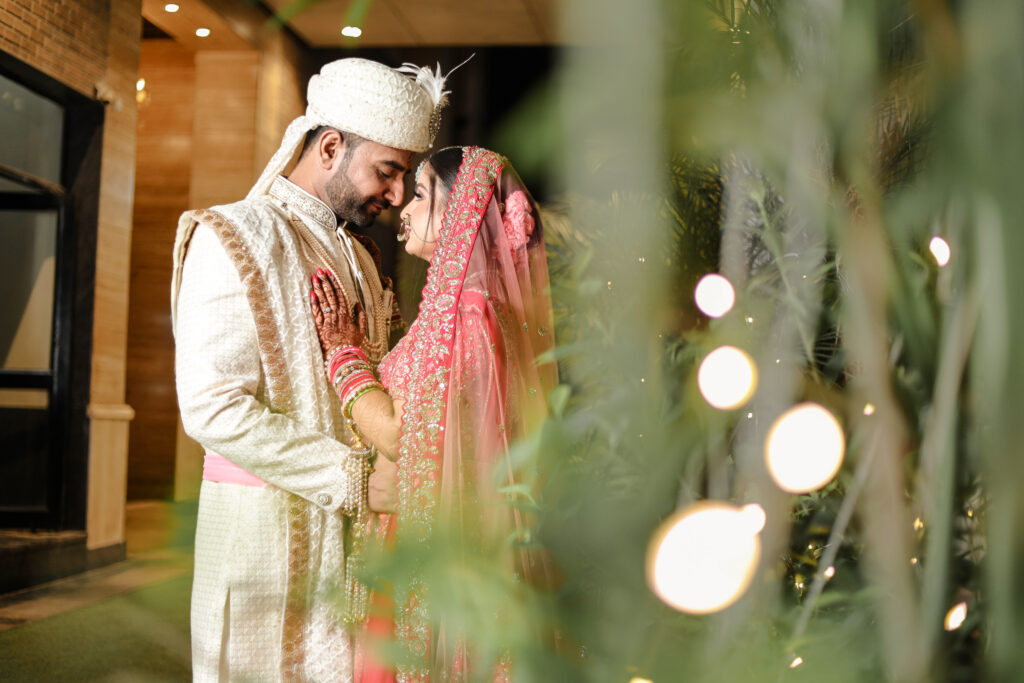 An intimate evening portrait of a newlywed couple standing close together and looking into each other's eyes, framed by softly blurred green foliage and glowing bokeh lights in the foreground. The groom wears a cream sherwani and turban, while the bride wears an ornate red lehenga and veil.