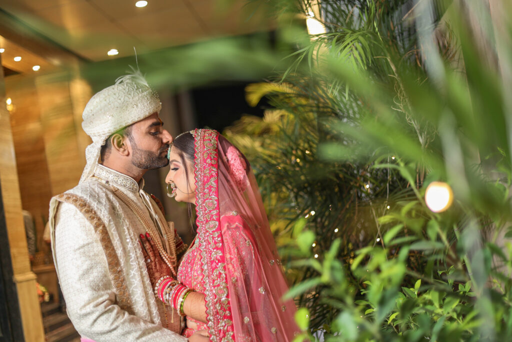 An intimate evening portrait of a newlywed couple close together, with the groom tenderly kissing the bride's forehead. The scene is softly framed by lush green foliage and bokeh lights in the foreground, with the groom wearing a cream sherwani and turban, and the bride wearing a traditional pink-and-red lehenga with a veil.