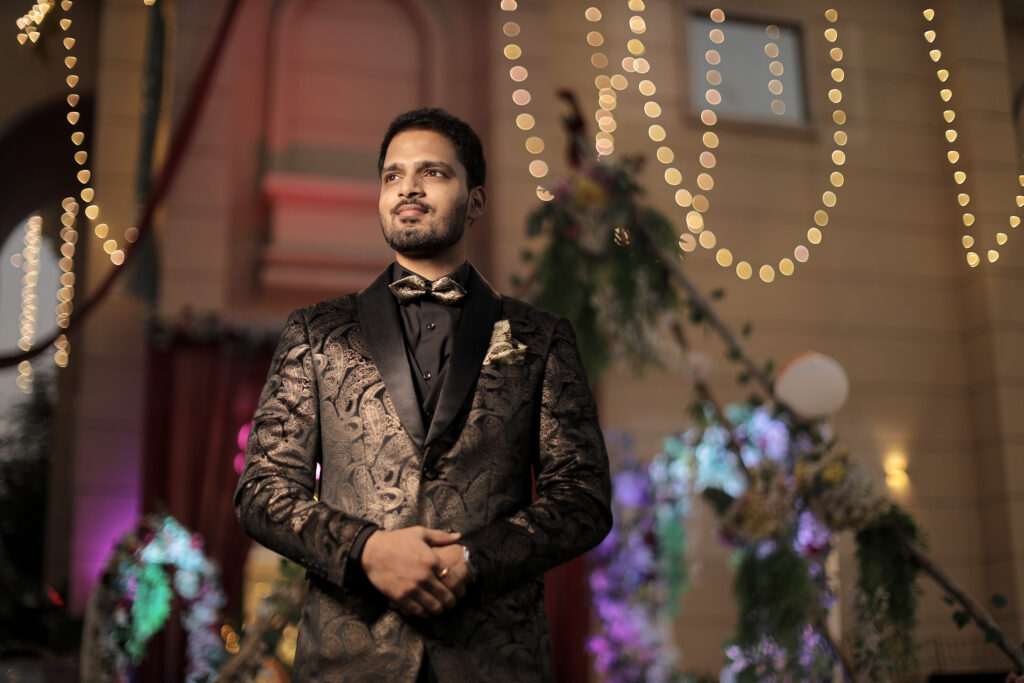 An outdoor portrait of a man standing proudly with his hands clasped, set against a festive venue background illuminated by cascading strings of warm fairy lights and floral arches. He wears a sophisticated black and gold patterned tuxedo jacket, a black shirt, and a matching bow tie.