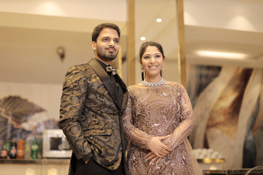 An indoor portrait of a couple standing side-by-side in a modern venue with a luxury interior background. The man wears a black and gold paisley-patterned tuxedo jacket with a bow tie, and the woman wears an embroidered rose-gold and mauve sheer gown with a sparkling necklace.