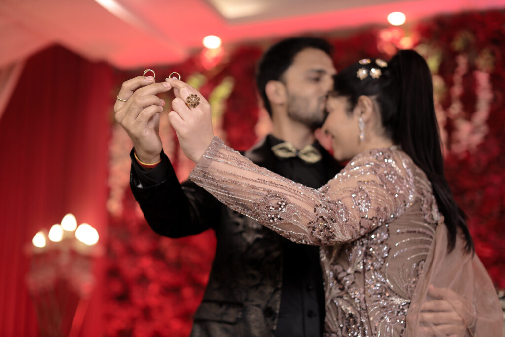An indoor portrait of a couple holding up their wedding bands with pride. The man gently kisses the woman's head while holding her in an embrace. The woman wears a glittering mauve-pink gown, and the man wears a dark patterned suit, set against a warm red floral backdrop.