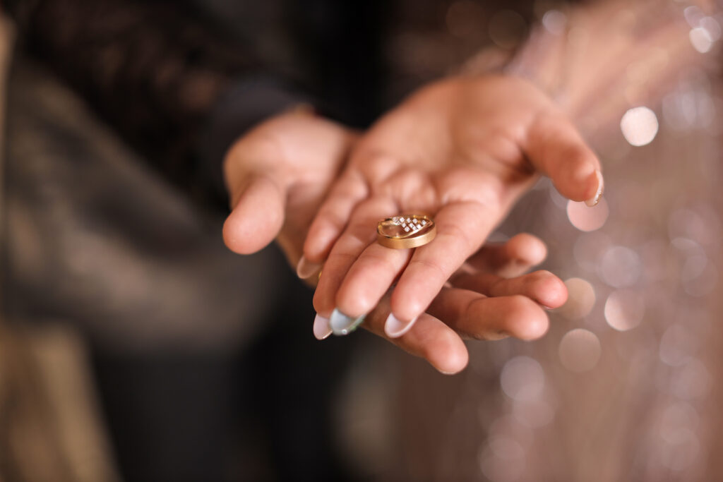 A close-up, macro shot of a couple's cupped hands holding a pair of nested gold wedding bands embellished with small sparkling diamonds, set against a softly blurred, warm bokeh background.