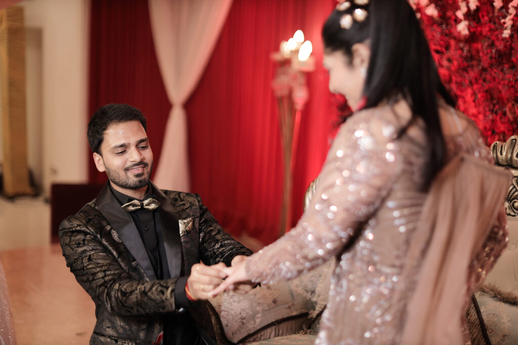 An indoor portrait capturing a man kneeling down to gently slide an engagement ring onto a woman's finger. The man looks up with a fond smile, wearing a dark patterned suit and bow tie. The woman stands facing away in a glittering mauve-pink gown, with her face softly blurred in the foreground.