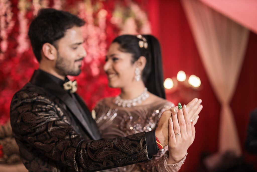 A close-up portrait of a couple holding hands and looking lovingly at each other with soft smiles. The man wears a dark patterned tuxedo and bow tie in the foreground, while the smiling woman is softly blurred in the background wearing a shimmering gown and a diamond necklace, set against a red floral backdrop.