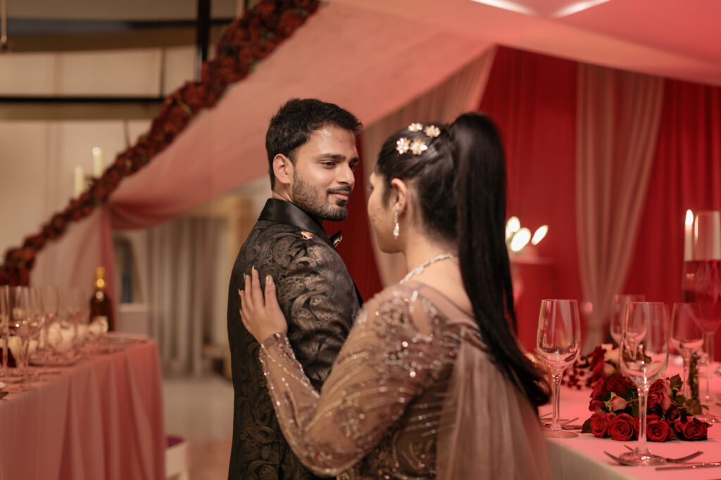 An indoor portrait of a couple standing close together in a warmly lit banquet hall decorated with red and pink tones. The man looks toward the camera in a dark patterned blazer, while the woman gently rests her hand on his shoulder, wearing a shimmering embroidered mauve-pink gown.