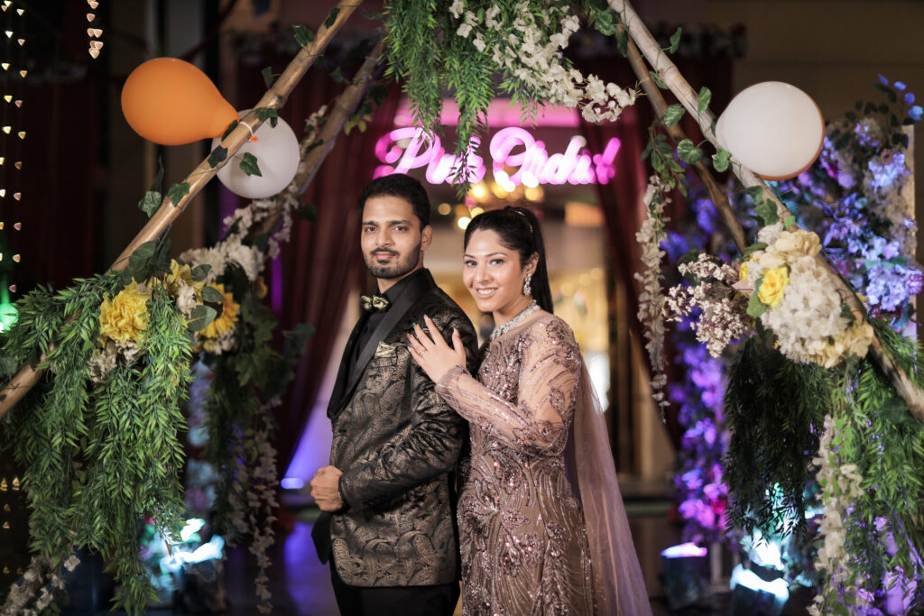 An indoor portrait of a couple standing beneath a rustic wooden archway decorated with lush green foliage, white and yellow flowers, and balloons. The man wears a patterned black and gold blazer and a bow tie, and the woman wears an embroidered mauve-pink gown with a sheer cape, placing her hand gently on his chest.