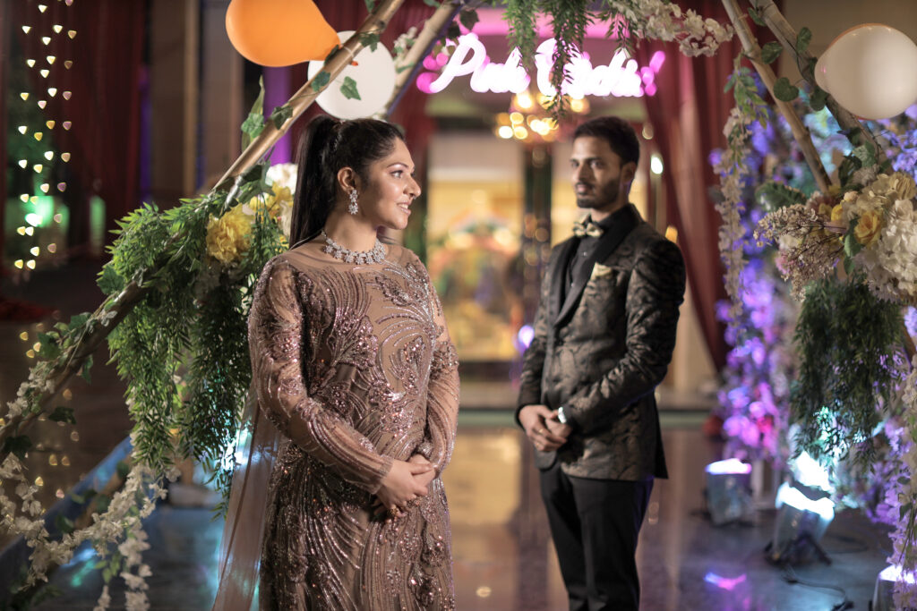 An indoor portrait featuring a woman in the foreground wearing a shimmering, embroidered mauve-pink gown and a sparkling necklace, looking off to the side. In the softly blurred background, a man stands beneath a rustic wooden arch decorated with greenery, white and yellow flowers, and balloons, wearing a dark patterned blazer and a bow tie.