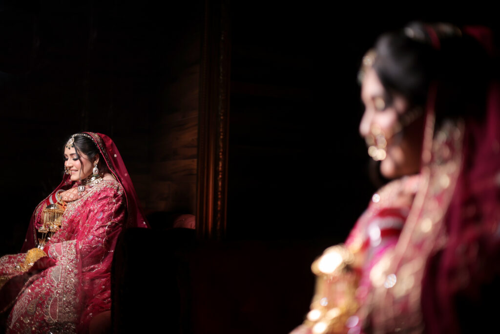An artistic indoor portrait of a bride dressed in a rich red embroidered lehenga and veil, sitting down and looking down gently. Her reflection is captured in a mirror, with a softly blurred view of her in the foreground against a dark background.