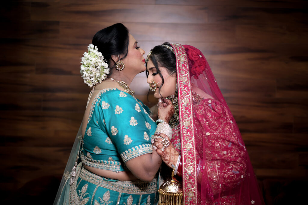 An intimate indoor portrait of an older woman in a teal and gold embroidered traditional outfit kissing the forehead of a bride dressed in a rich red lehenga and veil. They hold hands gently against a warm wooden background.