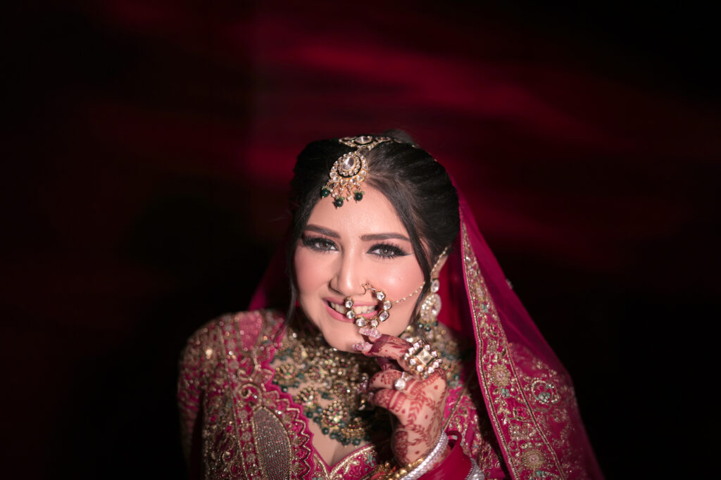A close-up indoor portrait of a smiling bride wearing a deep pink and gold embroidered lehenga and veil. She gently touches her traditional nose ring (nath) with a decorated hand, adorned with a maang tikka, statement necklace, and intricate mehendi.