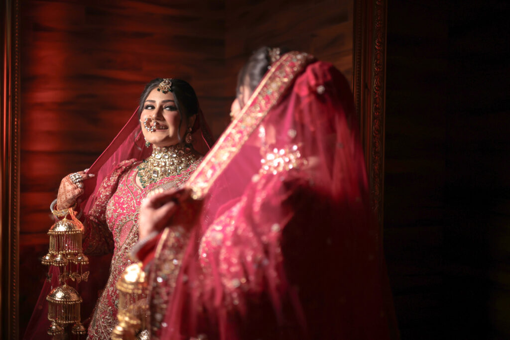 An indoor portrait of a smiling bride adjusting her red bridal veil, captured through a mirror reflection. Her back and side are softly blurred in the foreground, while her reflection clearly shows her wearing a richly embroidered pink-and-red lehenga, traditional kalire, a statement choker necklace, and a nose ring.