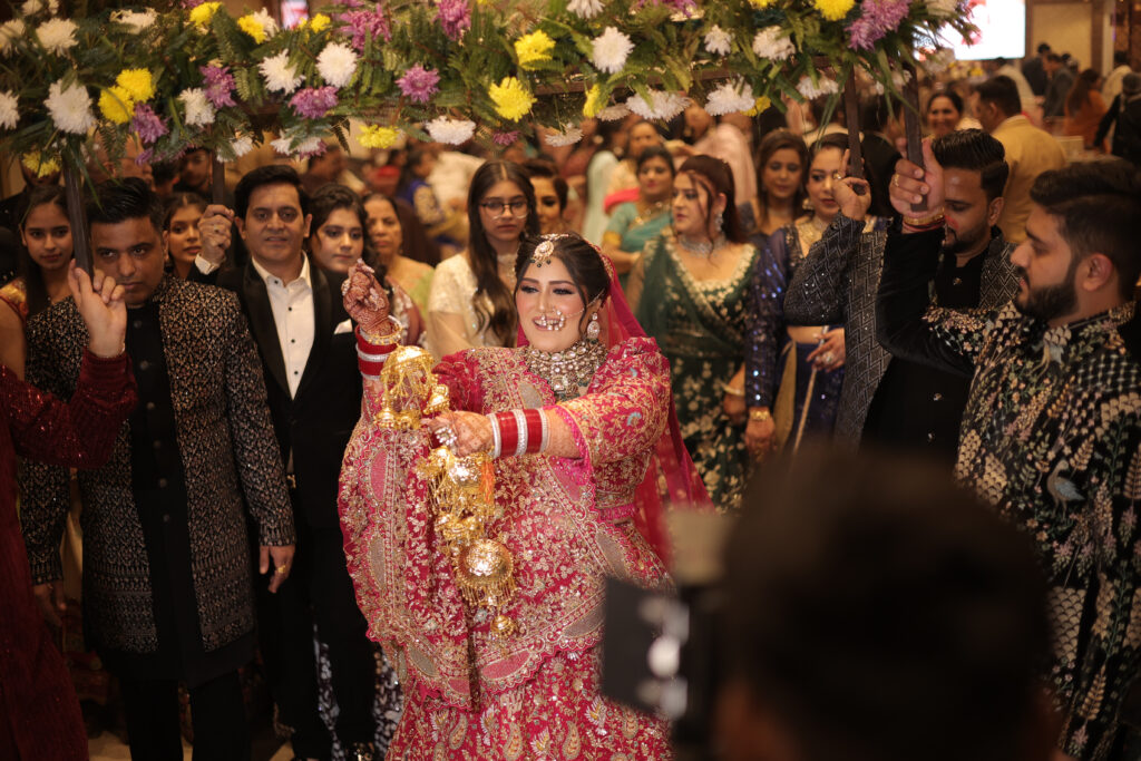 An indoor group portrait of a smiling bride in a vibrant pink and gold lehenga, performing a ritual while holding traditional kalire. She stands beneath a floral canopy (phoolon ki chaadar) held by family and friends during the wedding celebration.