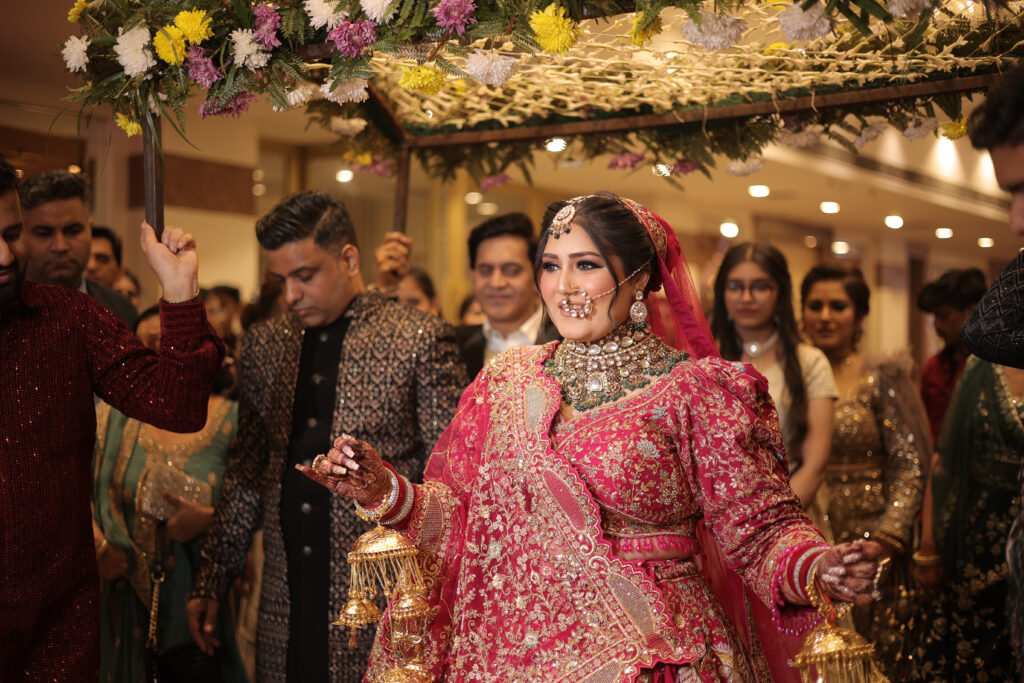 An indoor group portrait of a smiling bride walking underneath a decorated floral canopy (phoolon ki chaadar) held by family members during her wedding entry. She wears a vibrant pink and gold heavily-embellished lehenga, traditional kalire, a statement choker necklace, and a nose ring.