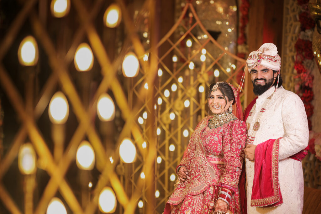 An indoor portrait of a newlywed couple smiling gently towards the camera, framed through a glowing lattice structure decorated with warm string lights. The bride wears a heavily-embellished red lehenga with traditional jewelry, while the groom stands beside her in a cream sherwani, turban, and a pink stole.