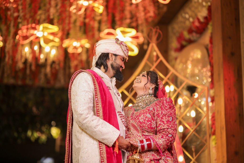 An indoor portrait of a newlywed couple smiling lovingly at each other, holding hands. The groom wears an embroidered cream sherwani, a turban, and a bright pink stole, while the bride shines in a detailed red lehenga, choker necklace, and traditional bridal bangles beneath a ceiling decorated with cascading floral arrangements and warm lights.