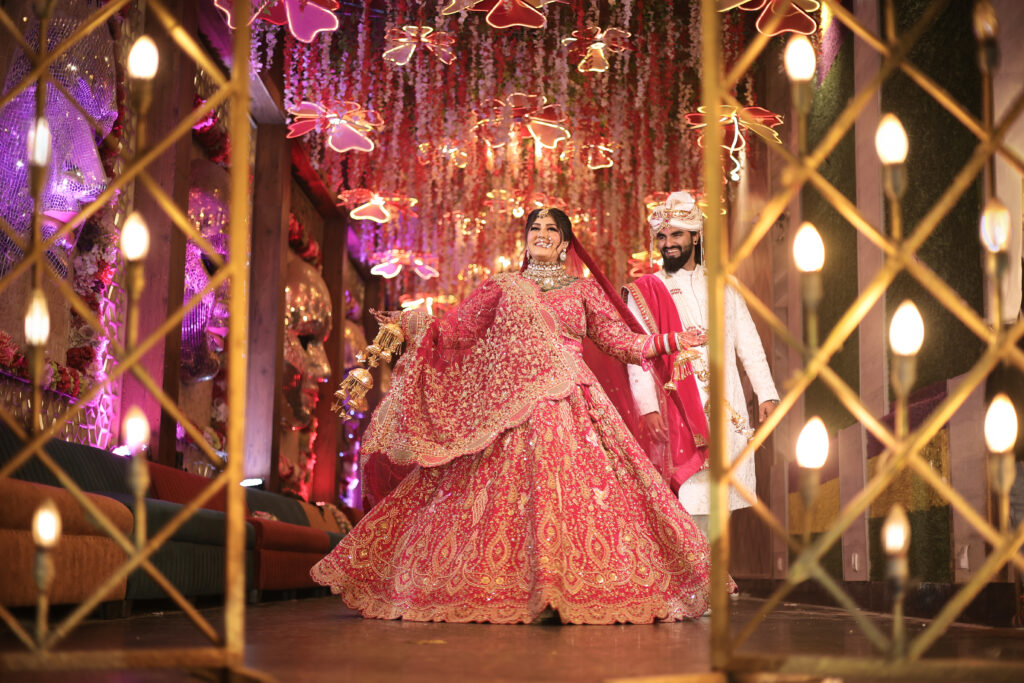 An indoor wide-angle portrait of a newlywed couple framed through a glowing, illuminated architectural archway. The bride twirls joyfully in a heavily-embellished red lehenga holding traditional kalire, while the groom stands nearby in a cream sherwani and turban, set against a corridor decorated with pink floral and light installations.