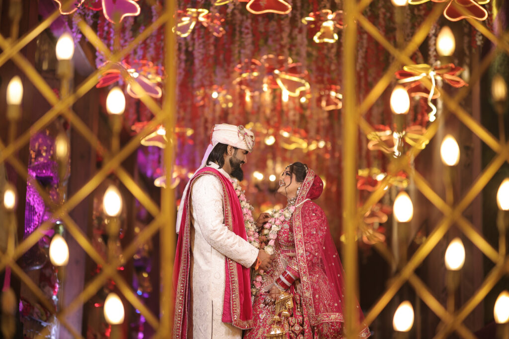 An indoor portrait of a newlywed couple smiling lovingly at each other, framed through an ornate golden lattice structure decorated with warm lights. The background features a ceiling filled with hanging flowers and illuminated flower-shaped fixtures.
