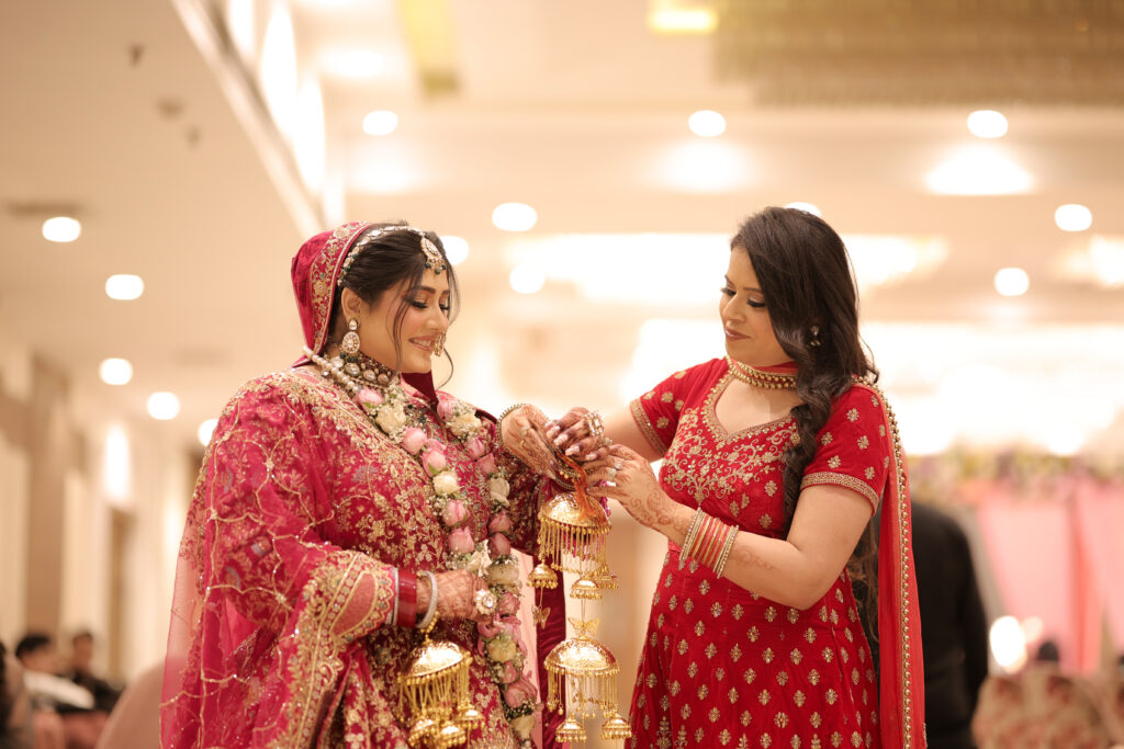 An indoor portrait of a bride in a rich red lehenga and floral garlands smiling as a family member helps her with the traditional kalire ritual. The family member wears a matching red embroidered suit.