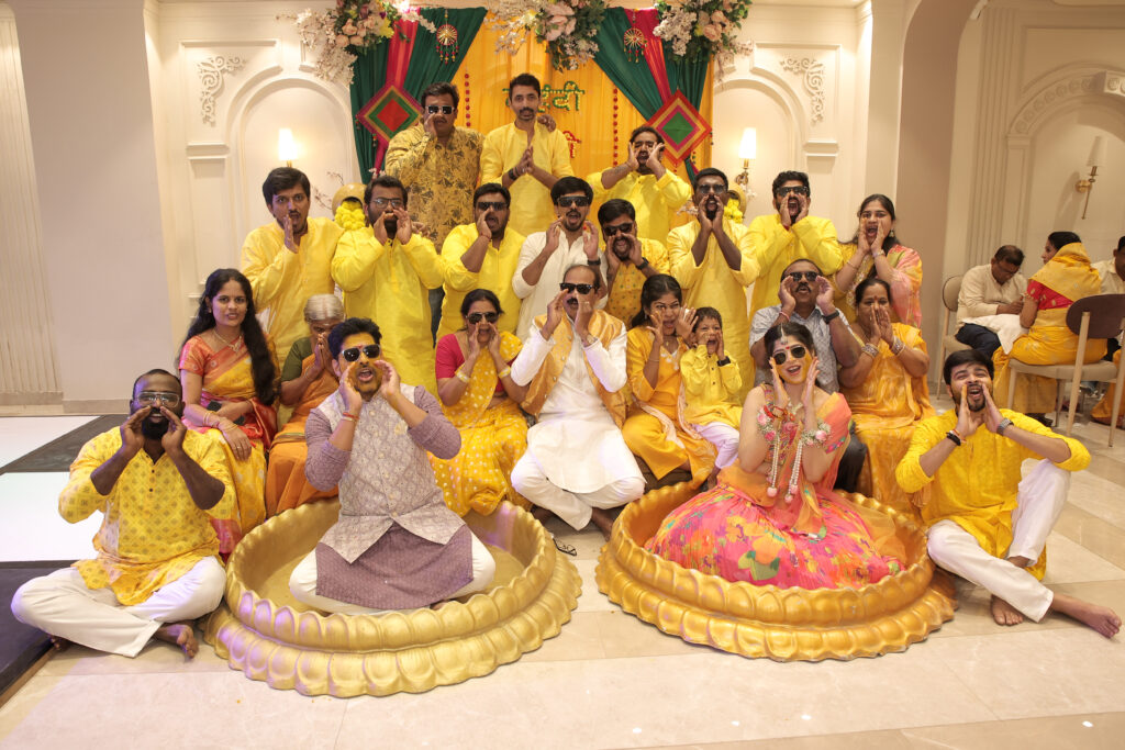 A wide indoor group portrait of family and friends celebrating a Haldi ceremony, with two people seated in golden lotus-shaped basins. Everyone wears yellow or traditional attire and sunglasses, striking playful poses with hands cupped around their mouths.