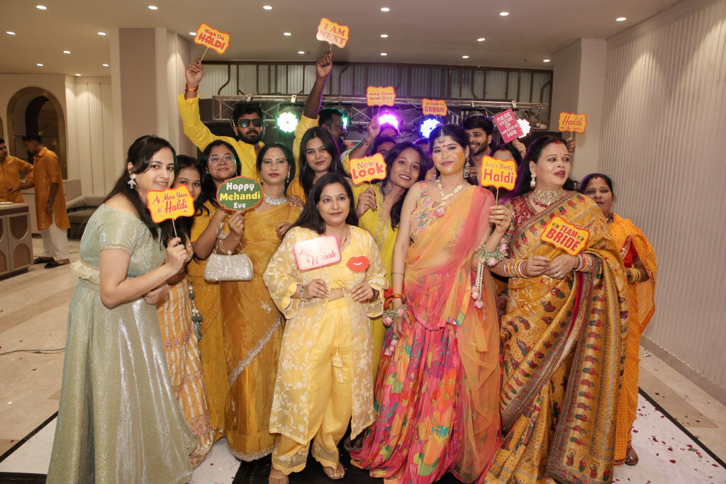 A wide indoor group portrait of a bride and her family and friends holding colorful celebratory props and signs. The bride wears a vibrant pink-and-yellow lehenga, while guests around her wear shades of yellow, green, and traditional ethnic attire, smiling happily together.
