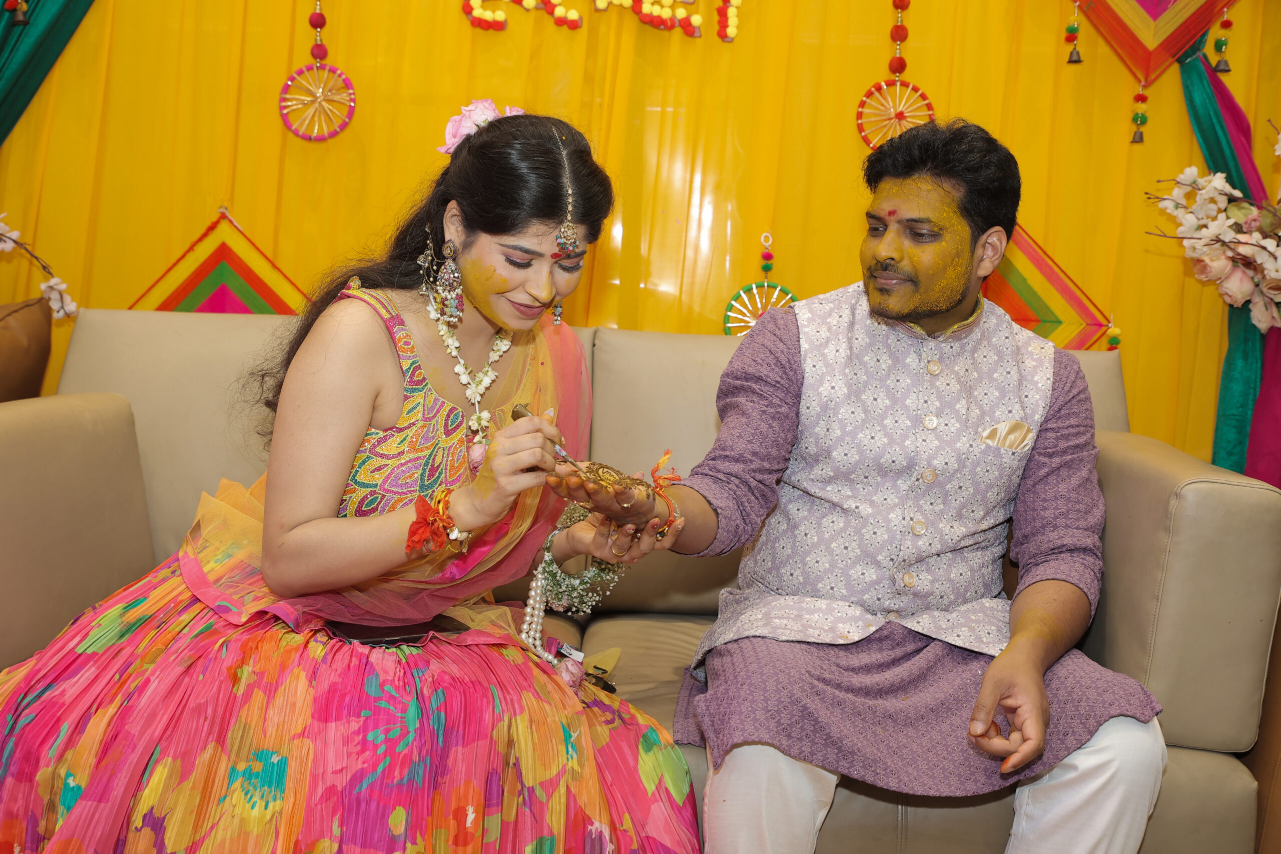 An indoor portrait of a couple participating in a traditional Haldi ceremony, seated together on a light sofa against a bright yellow decorated background. The woman wears a colorful floral lehenga and has haldi on her face, while applying a ritualistic element to the man, who wears a purple kurta and an embroidered grey Nehru jacket with haldi on his face.
