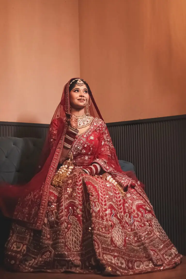 A seated indoor portrait of a smiling Indian bride looking upward off-camera. She is wearing a heavily embroidered red bridal lehenga featuring peacock motifs, a sheer red dupatta draped over her head, and traditional bridal jewelry including a large nath, choker, and kalire.